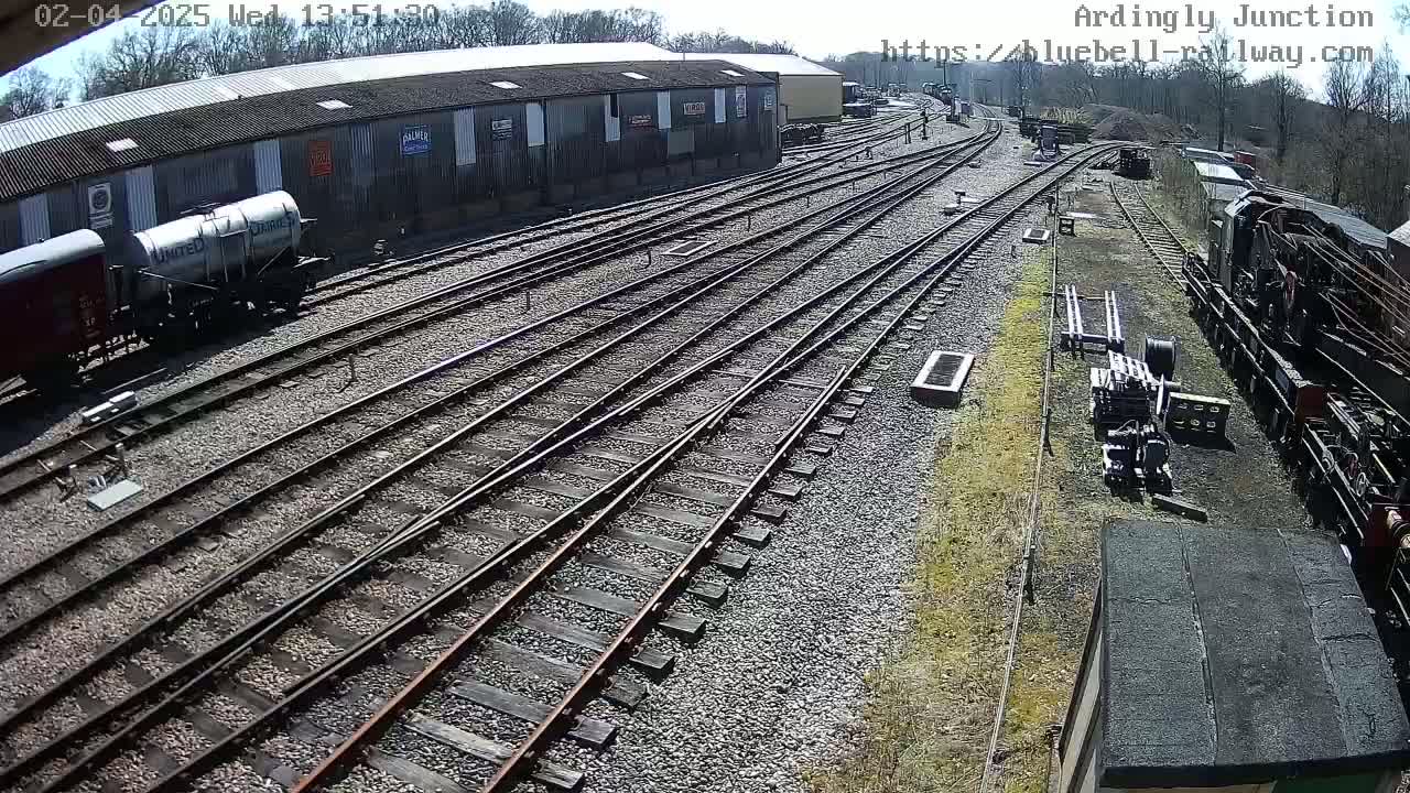 A railway yard with multiple train tracks, several train cars, and a large building under a sunny sky.