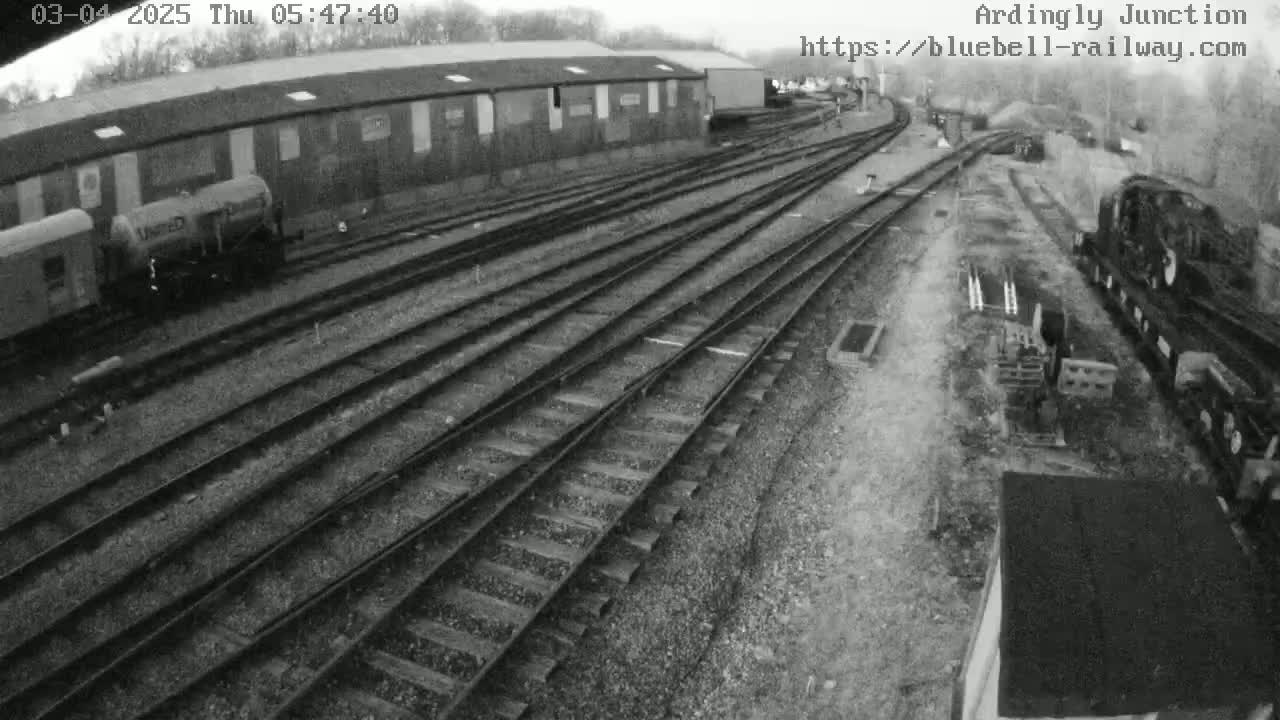 A high-angle, grayscale view of a railway yard with multiple train tracks, railway cars, and a building, on an overcast day.