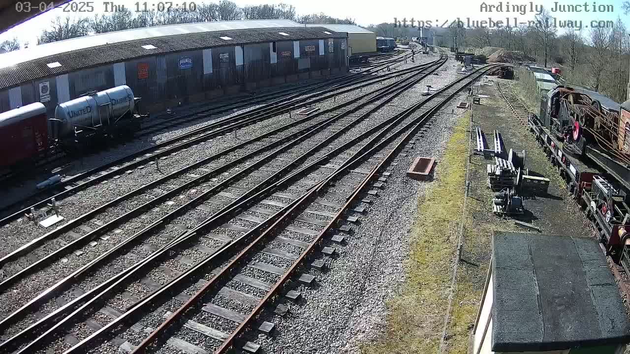 A sunny day at a railway yard shows multiple train tracks branching out from a central point, with several railcars and locomotives parked nearby.