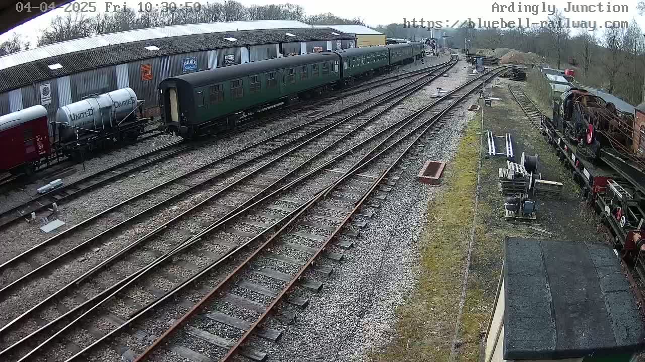 A railway yard with several railcars and locomotives, some parked alongside a long building, on an overcast day.
