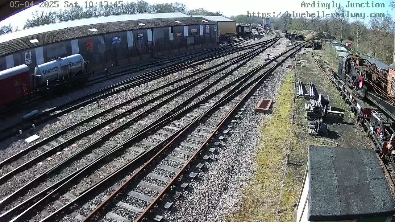 A sunny day at a railway yard shows multiple train tracks branching out from a large building with rail cars and locomotives nearby.