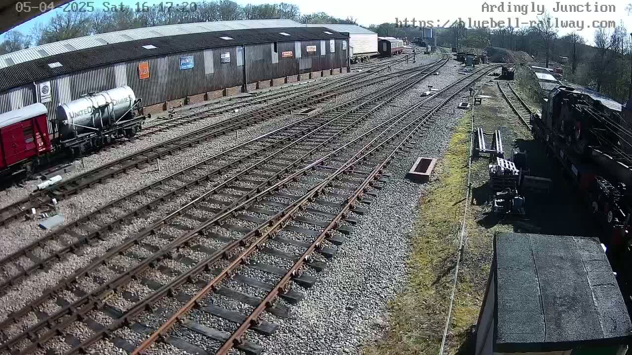 A sunny day at a railway yard shows multiple train tracks branching out from a long building with railcars and locomotives parked nearby.