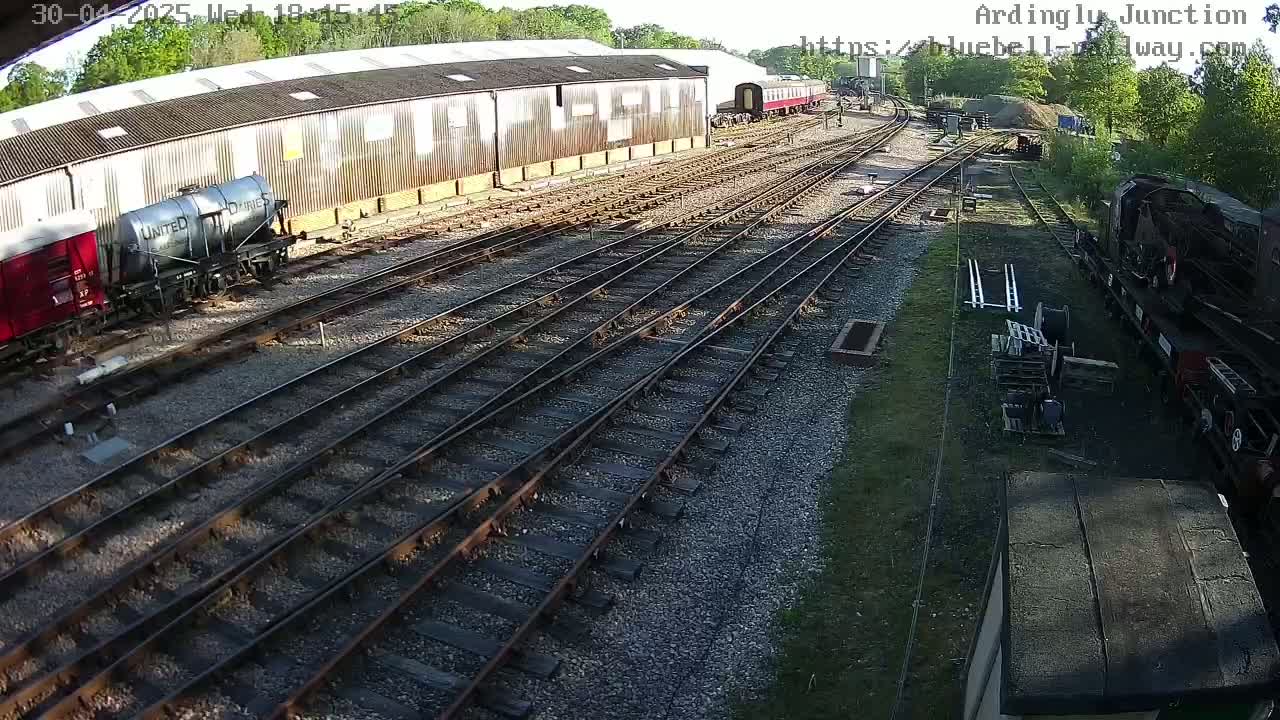 A railway yard with multiple train cars, including a tanker car, parked next to a long building on a sunny day.