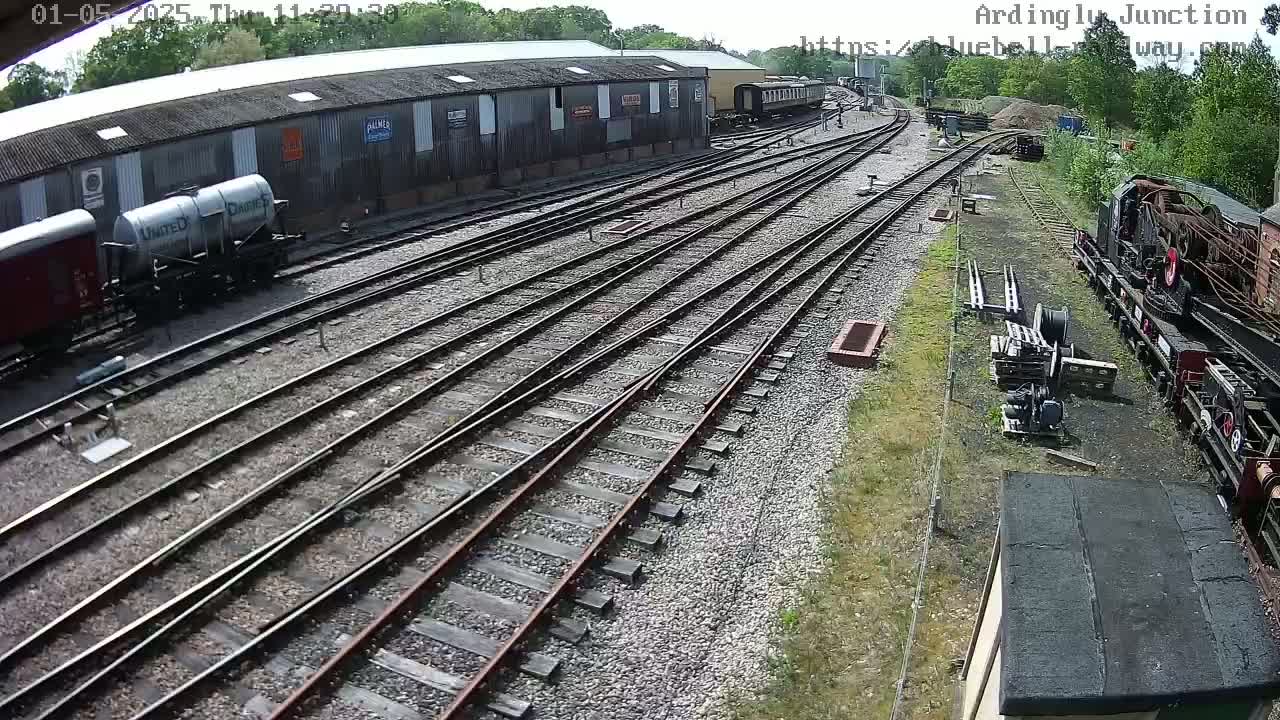 A railway yard with multiple train tracks, several railway cars, and a large shed under a sunny sky.