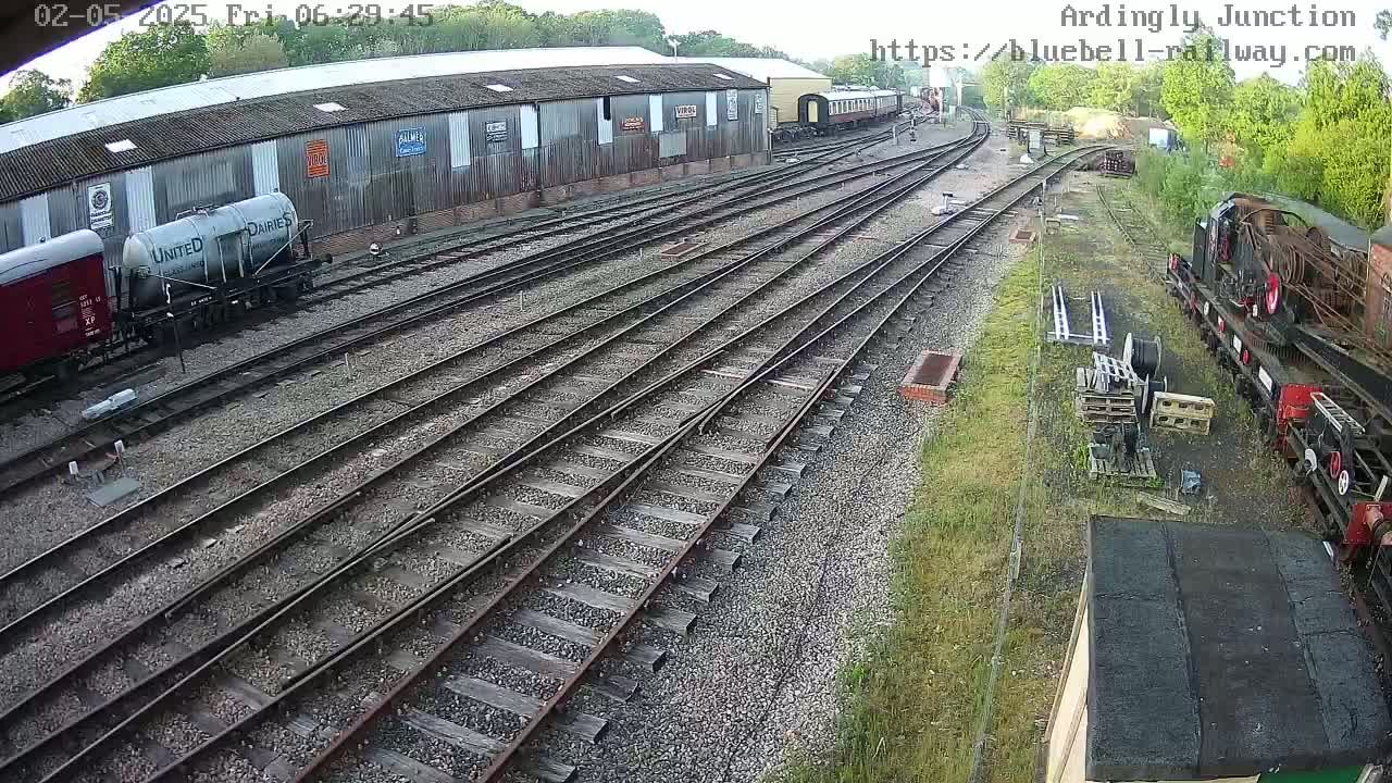 Several railway cars and locomotives are parked on a sprawling network of train tracks next to a long, gray building, on a partly sunny day.