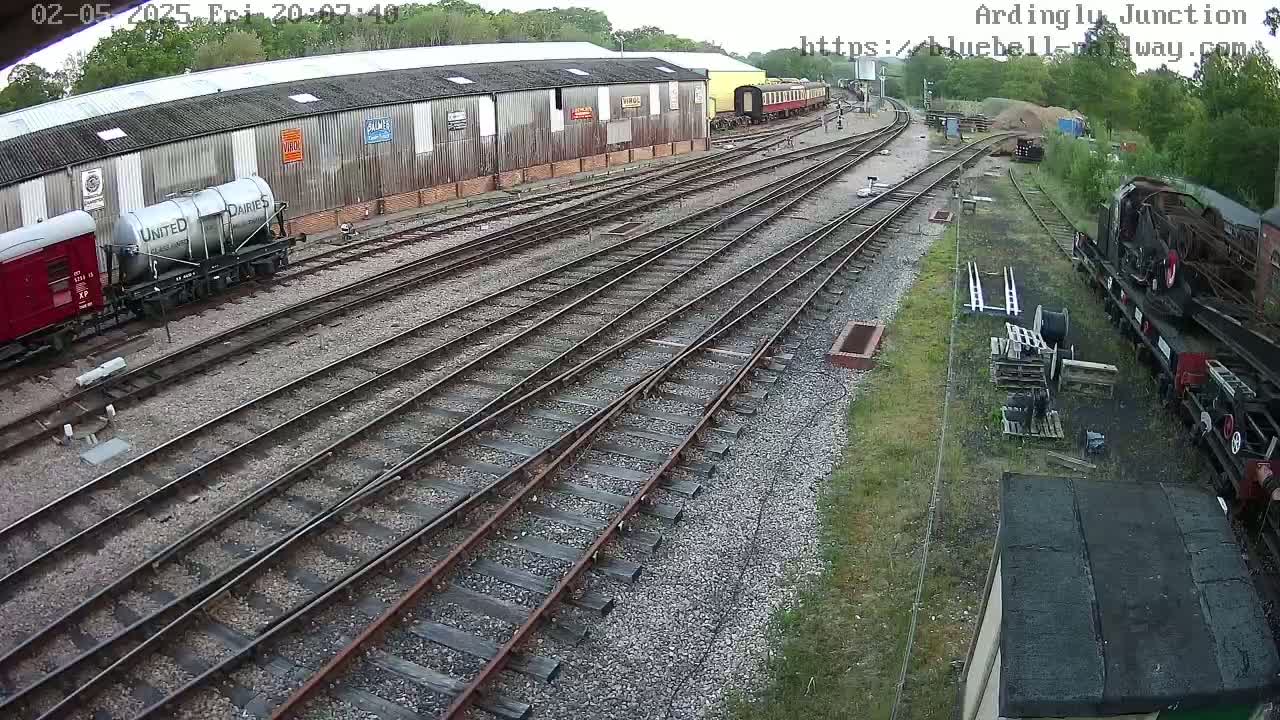 A high-angle view shows a railway yard with multiple train cars, rail lines, and a large wooden building, under an overcast sky.