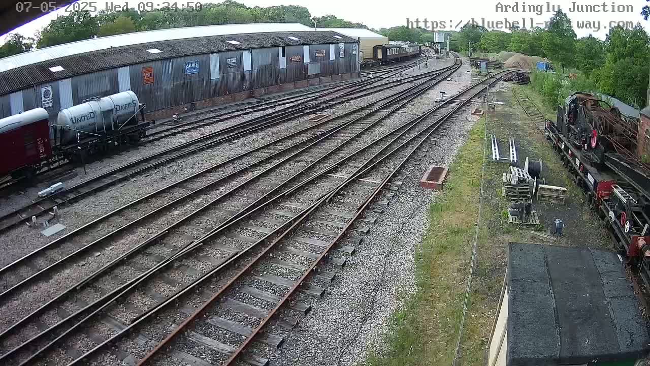 A railway yard with multiple train cars, a long shed, and several sets of railway tracks, on a partly sunny day.