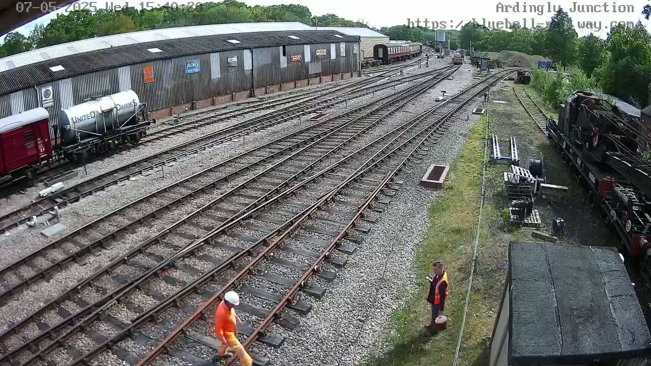 A high-angle view shows a railway yard with multiple train tracks, several railway cars, and two people in orange vests working near the tracks on a partly sunny day.