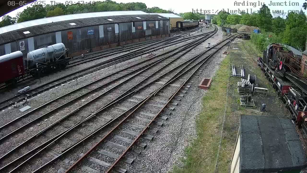 Several railway cars are parked near a long building alongside multiple sets of train tracks on a mostly sunny day.