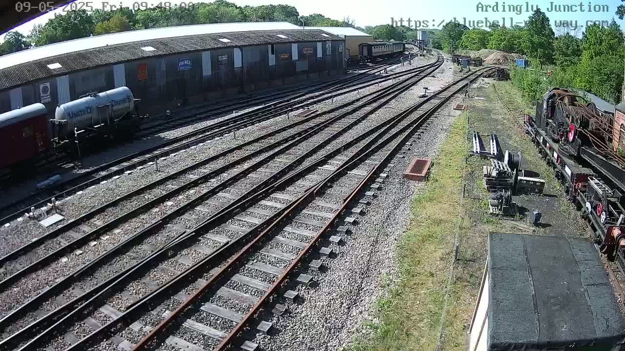 A sunny day at a railway yard shows multiple train cars, tracks, and a building.