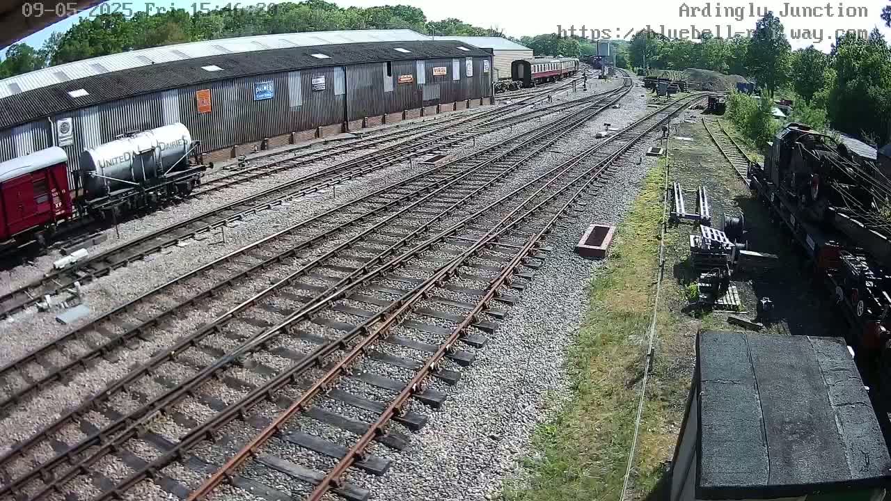 A sunny day at a railway yard shows multiple train tracks branching out from a large building, with several railway cars and a partially visible locomotive present.