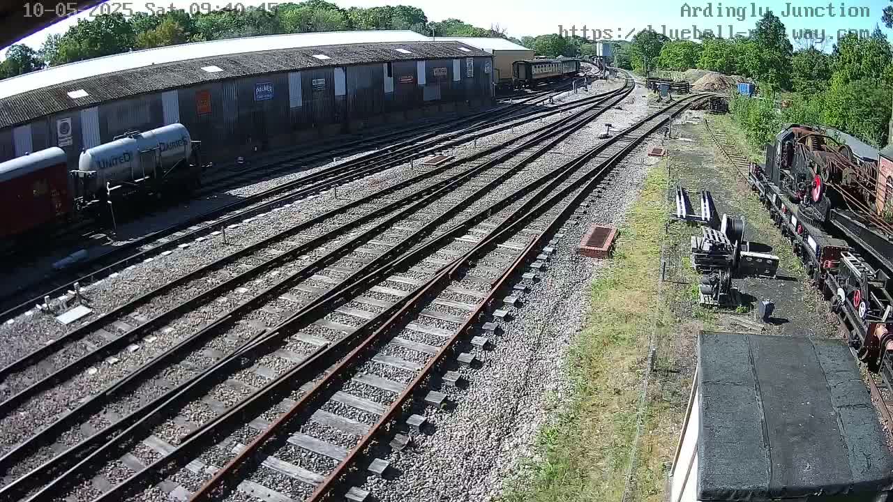 A sunny day at a railway yard shows multiple train tracks branching off from a large building with several railcars parked nearby.