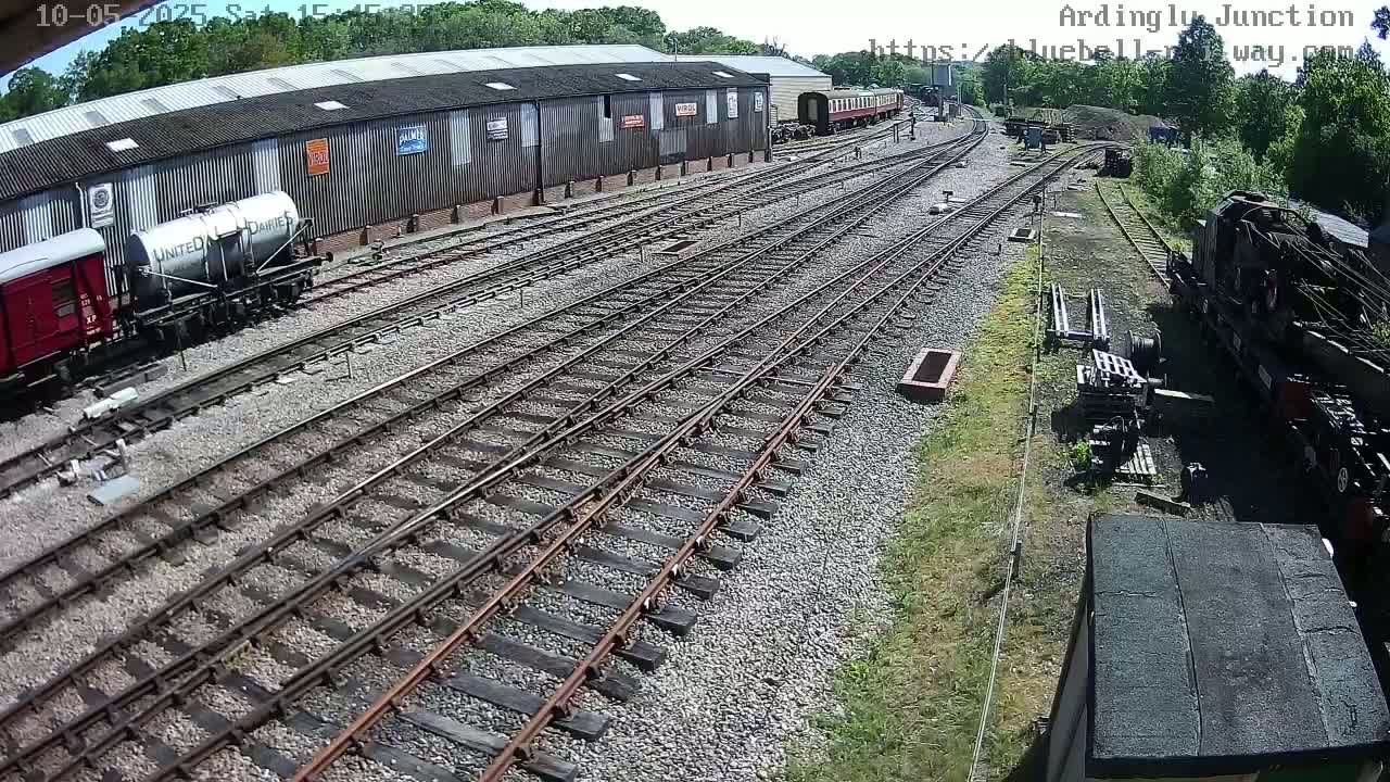 A sunny day at a railway yard shows multiple train tracks branching off from a main line, with several railcars and a locomotive parked nearby.