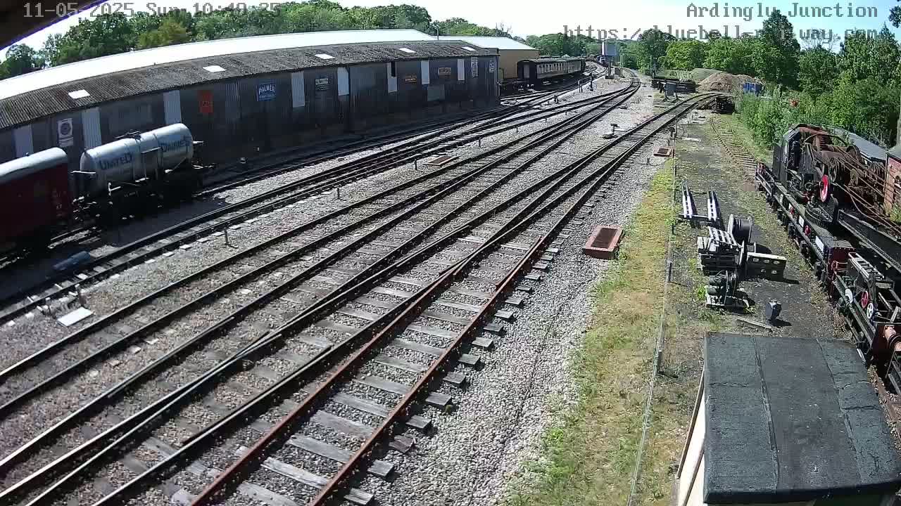 A sunny day at a railway yard shows multiple train tracks branching out from a long shed with rail cars parked nearby.