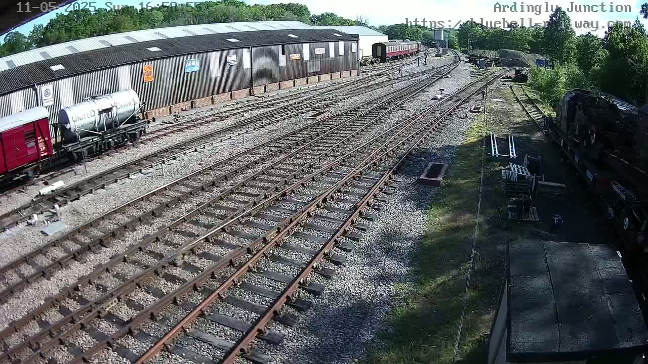 A sunny day at a railway yard shows multiple train cars and tracks, including a red boxcar, a tanker car, and a partially visible locomotive.