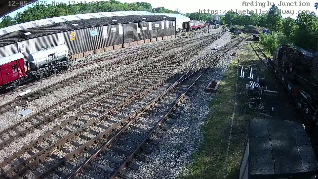A railway yard with multiple train cars and tracks, partially shaded by trees on a sunny day.