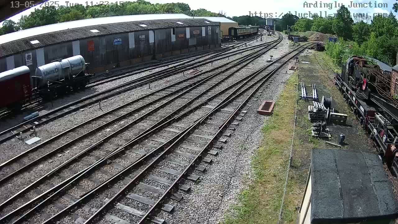A sunny day at a railway yard shows multiple train cars and locomotives near a long building and intersecting train tracks.