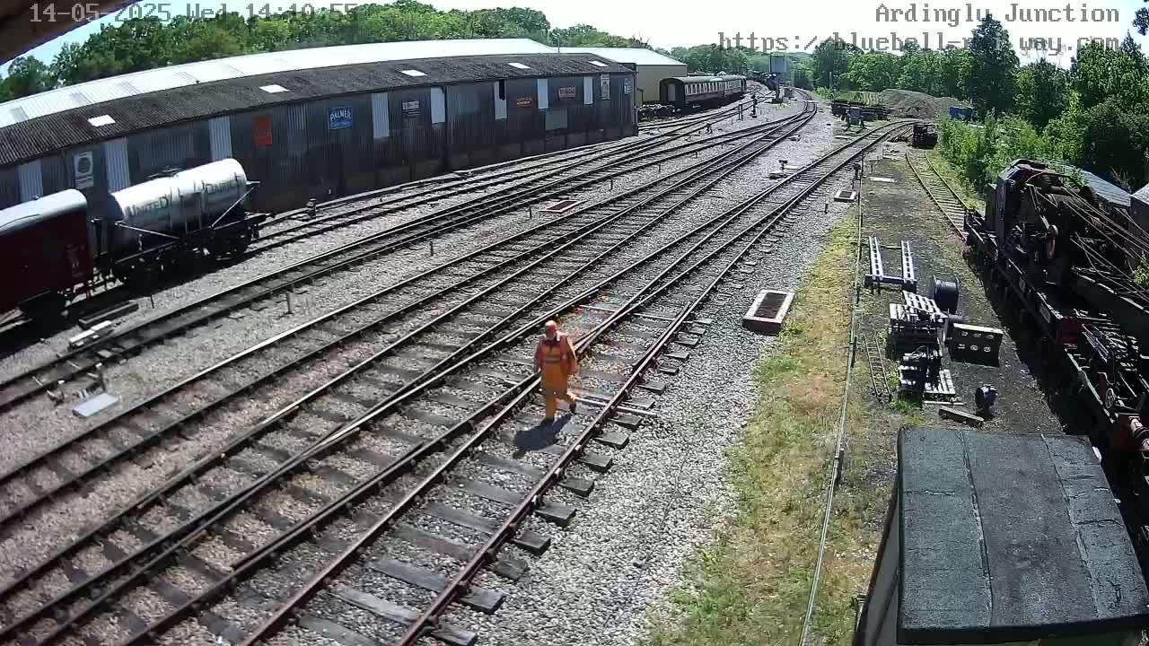 A person in orange walks across railroad tracks next to a train yard on a sunny day.