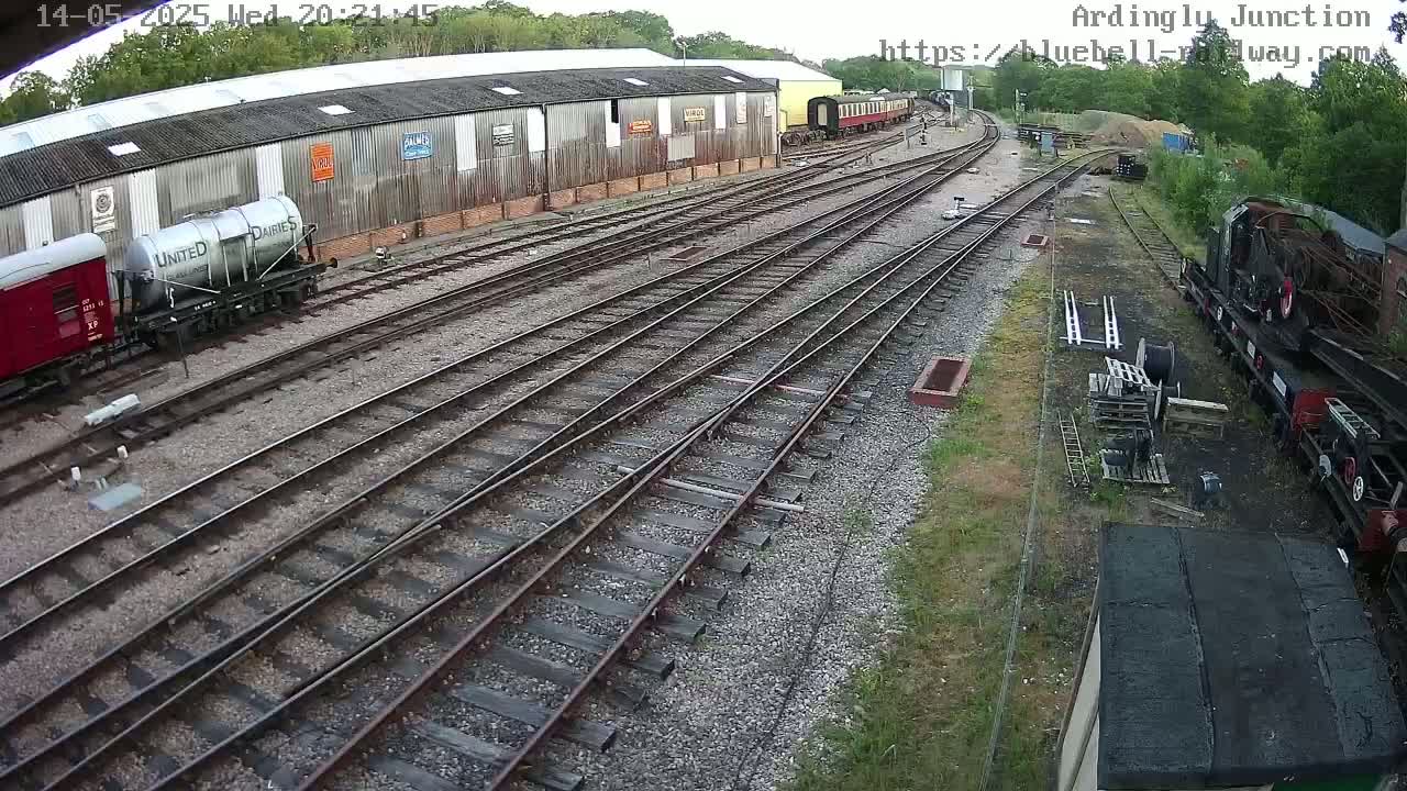 A railway yard with several train cars and a long warehouse is viewed from above on a partly sunny day.