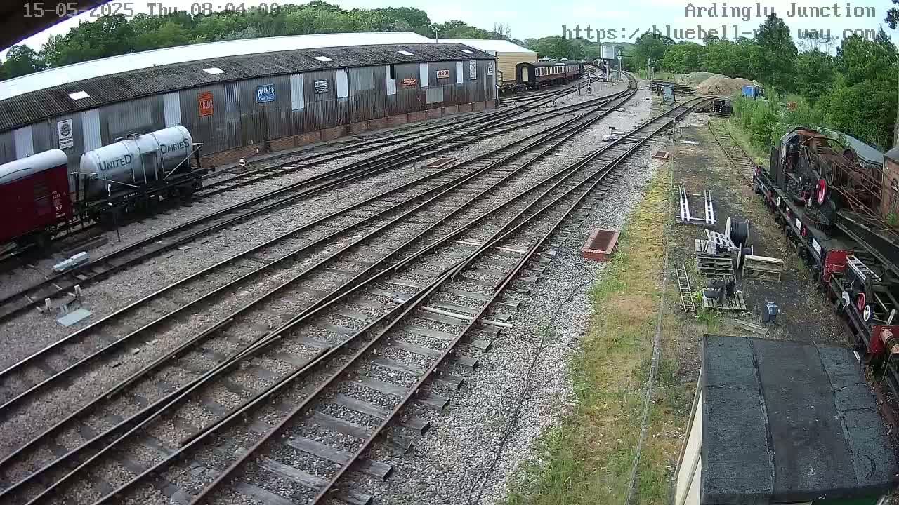 A railway yard with multiple train cars, some parked and others on tracks, is shown on a partly sunny day.