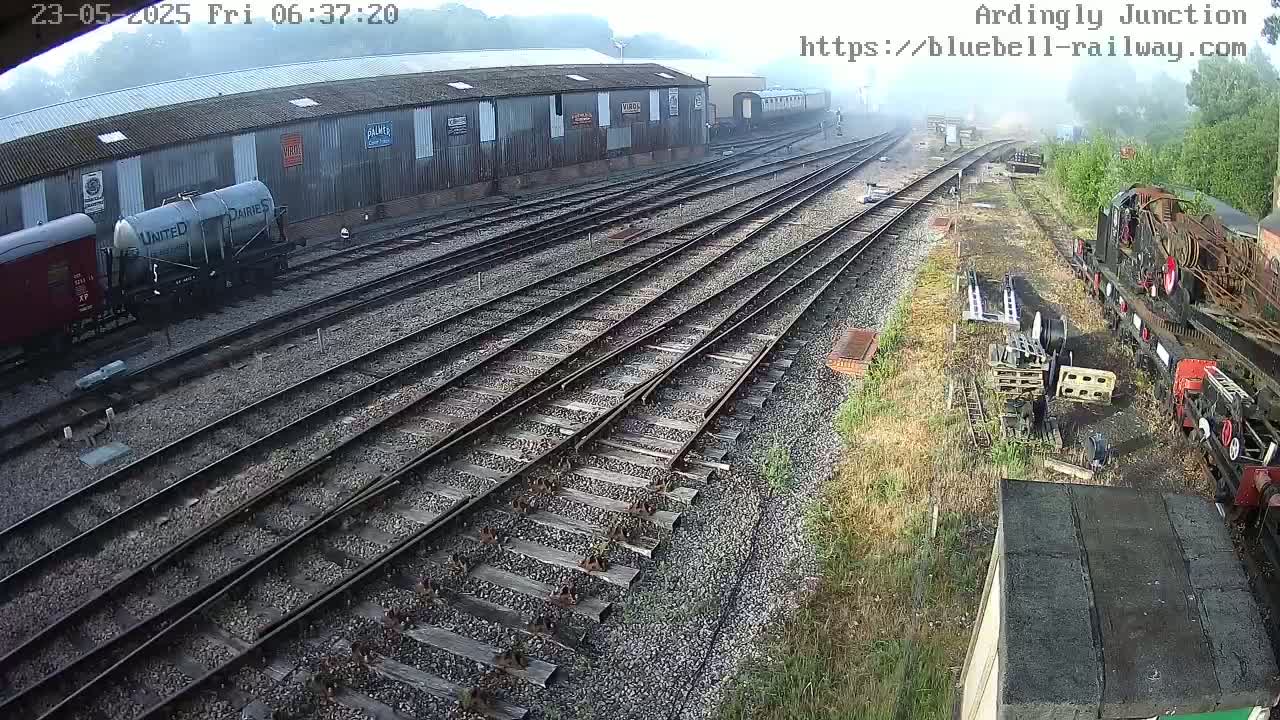 A high-angle view of a railway yard with multiple train tracks, railway cars, and a partially visible steam locomotive on a misty morning.