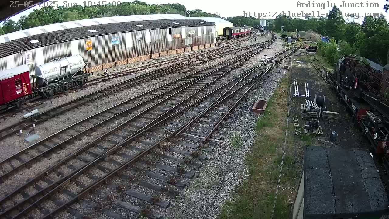 A railway yard with multiple train tracks, railcars, and a long warehouse, under a partly cloudy sky.