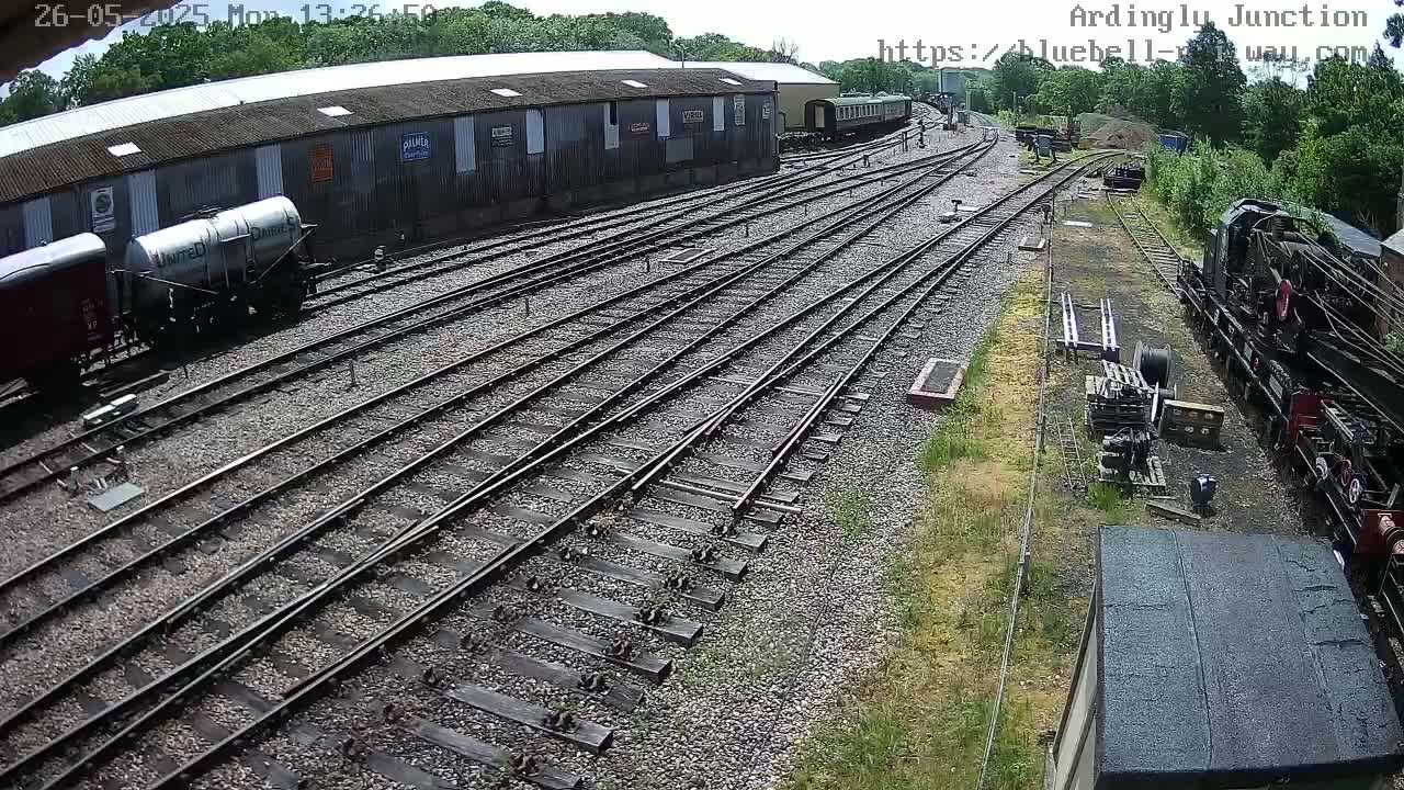 A high-angle view shows a railway yard with multiple train tracks, several rail cars, and a partially visible locomotive under a sunny sky.