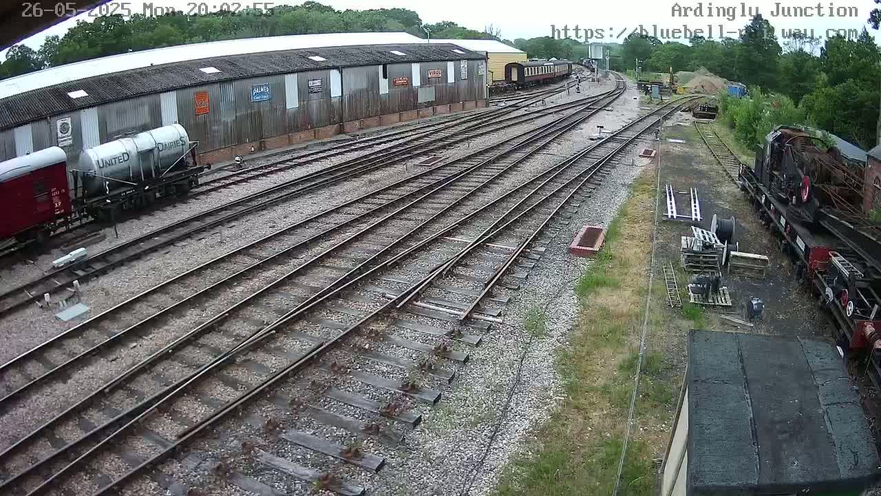 A high-angle view shows a railway yard with multiple train tracks, several rail cars, and a partially visible locomotive under a sunny sky.