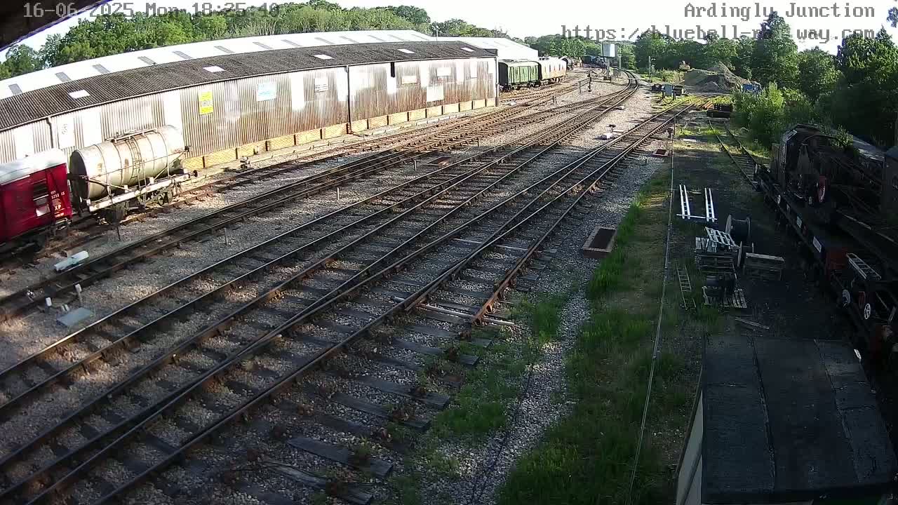 A high-angle view shows a railway yard with multiple train cars parked near a long shed on a sunny day.