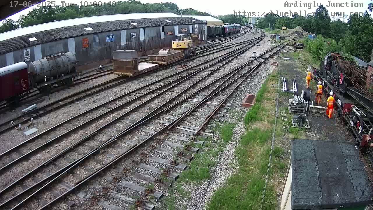 A railway yard with multiple train cars, a small excavator, and several workers in orange safety vests under a partly cloudy sky.