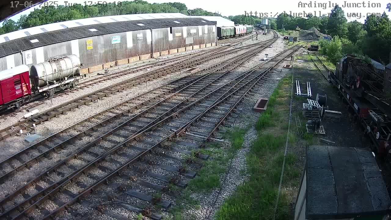 A high-angle view shows a railway yard with multiple train cars and tracks on a sunny day, alongside a long building.