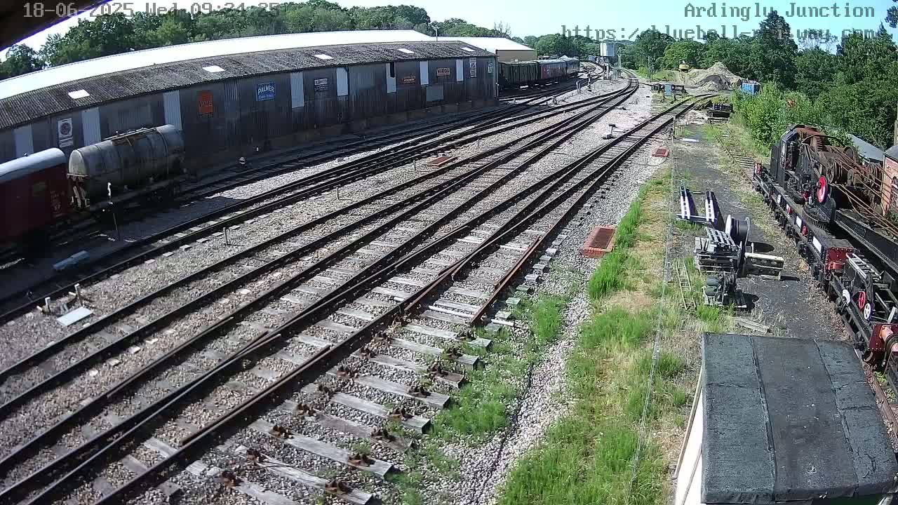 A sunny day at a railway yard shows multiple train tracks branching out from a long building with railway cars parked alongside it, and a train partially visible in the lower right.