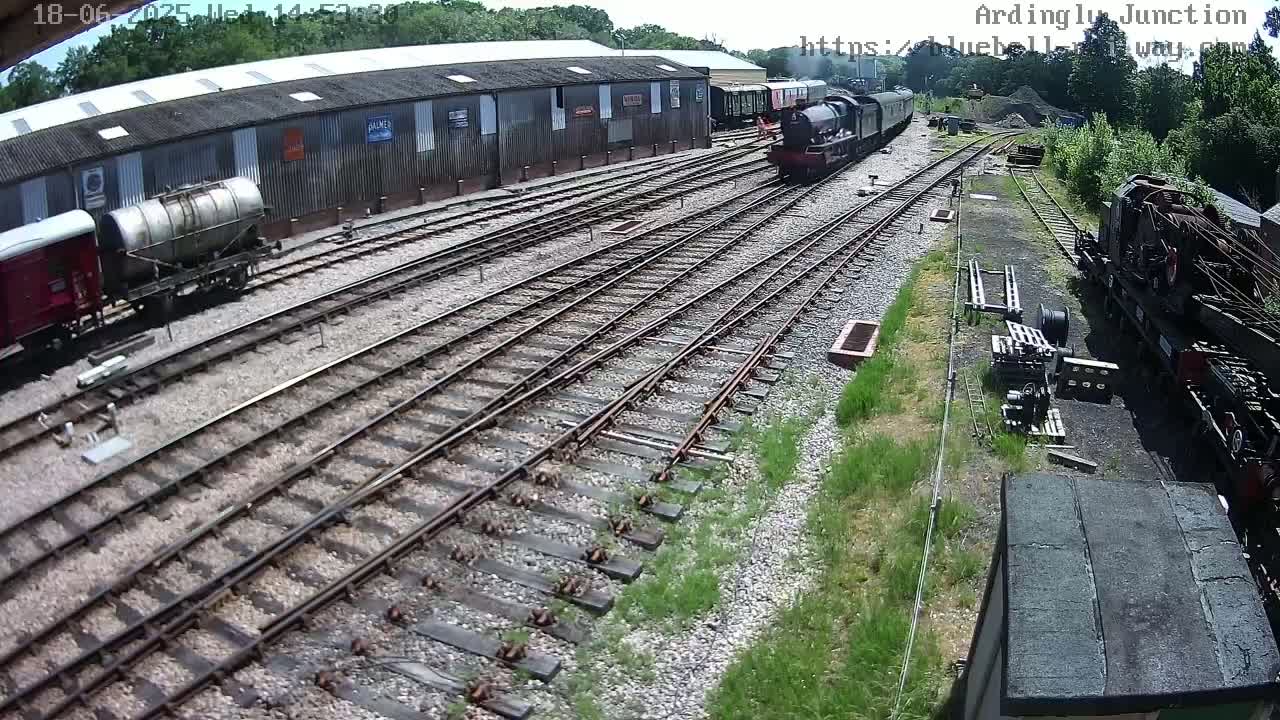 A high-angle view of a railway yard on a sunny day, showing several train cars and locomotives near a long building, with multiple tracks stretching into the distance.
