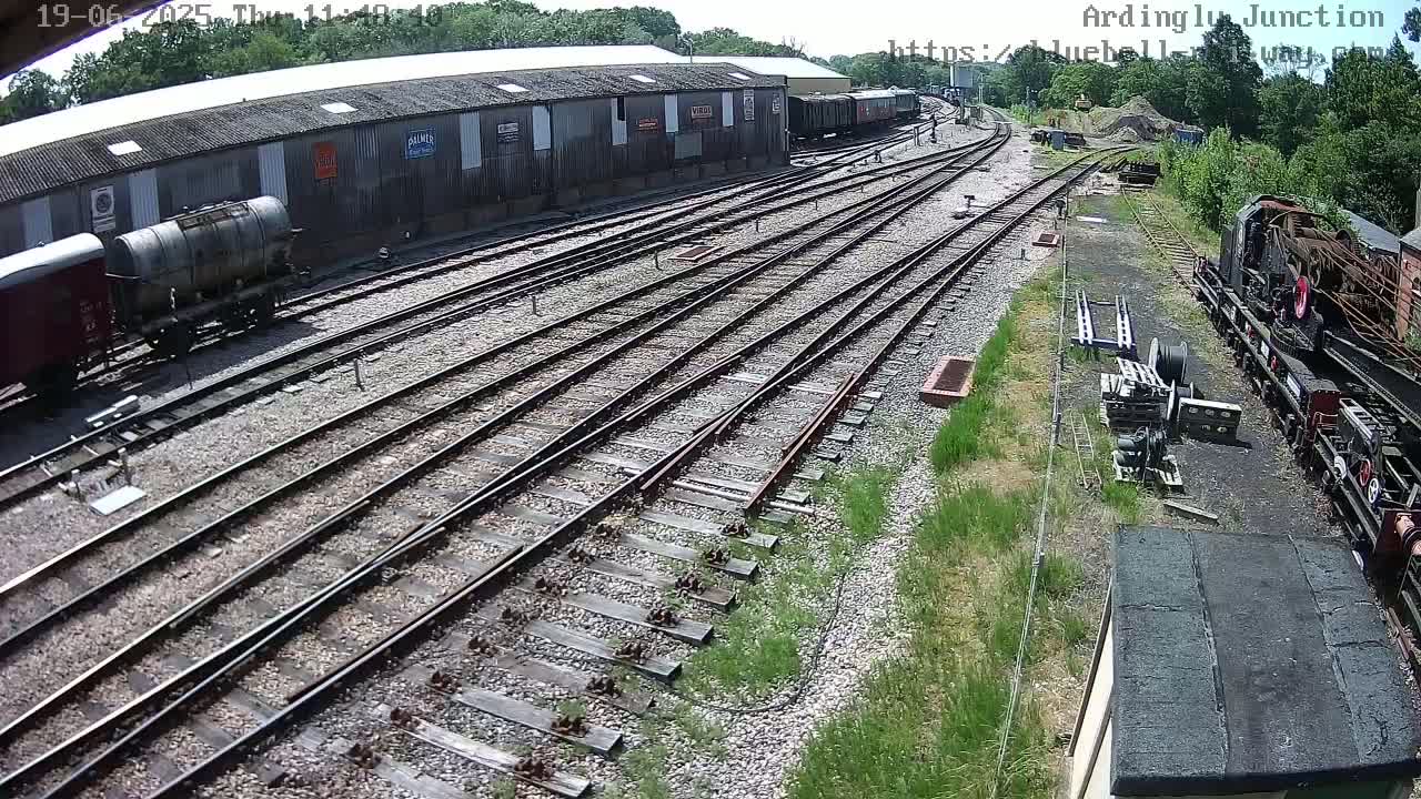 A high-angle view of a railway yard on a sunny day shows numerous train tracks branching out from a long building with several railway cars and locomotives nearby.