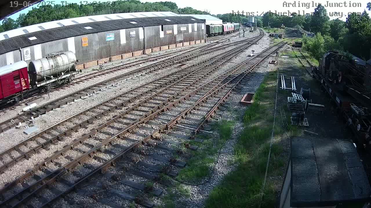 A sunny day at a railway yard shows multiple sets of train tracks intersecting, with several train cars and a large warehouse in the background.