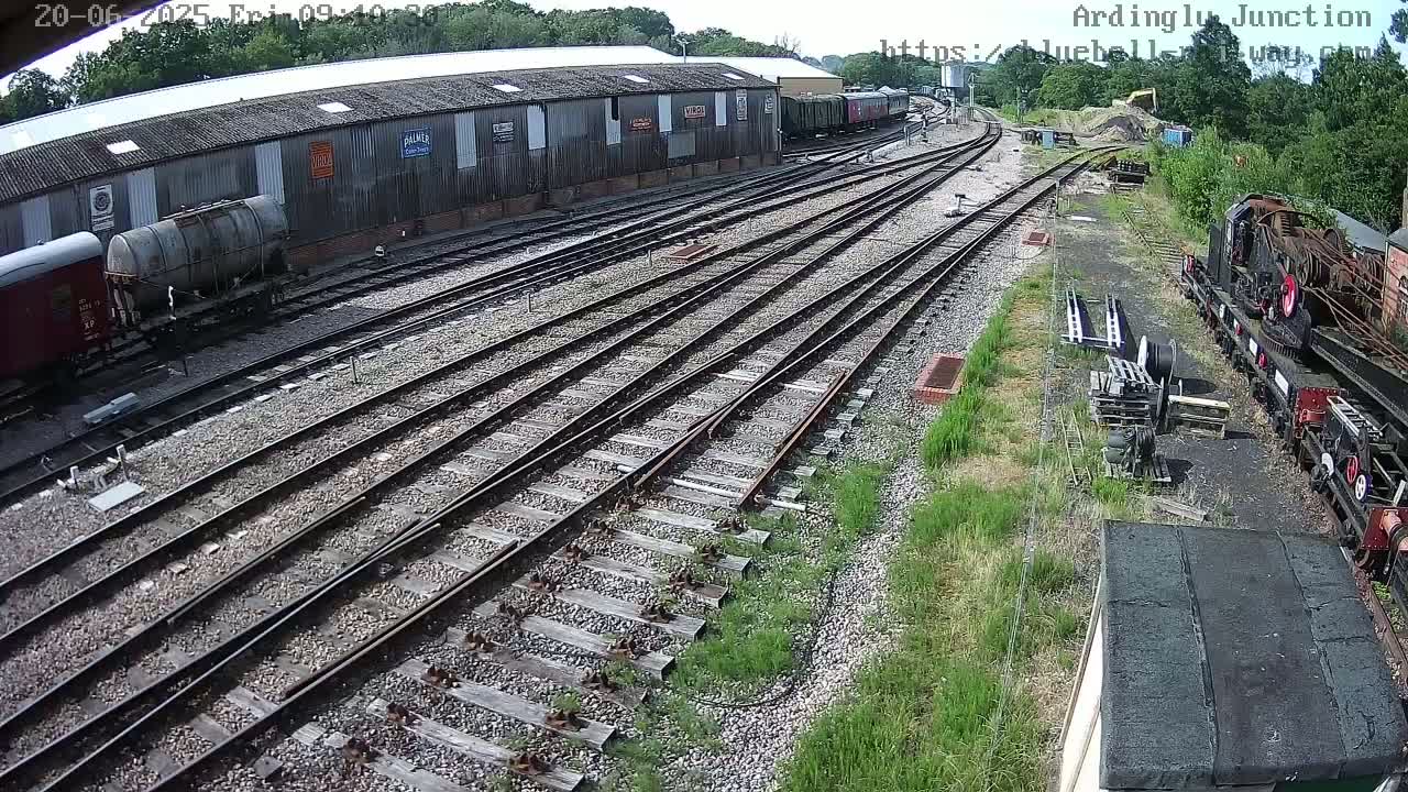 A high-angle view of a railway yard on a sunny day shows numerous train tracks branching out from a large building with several railway cars and locomotives nearby.