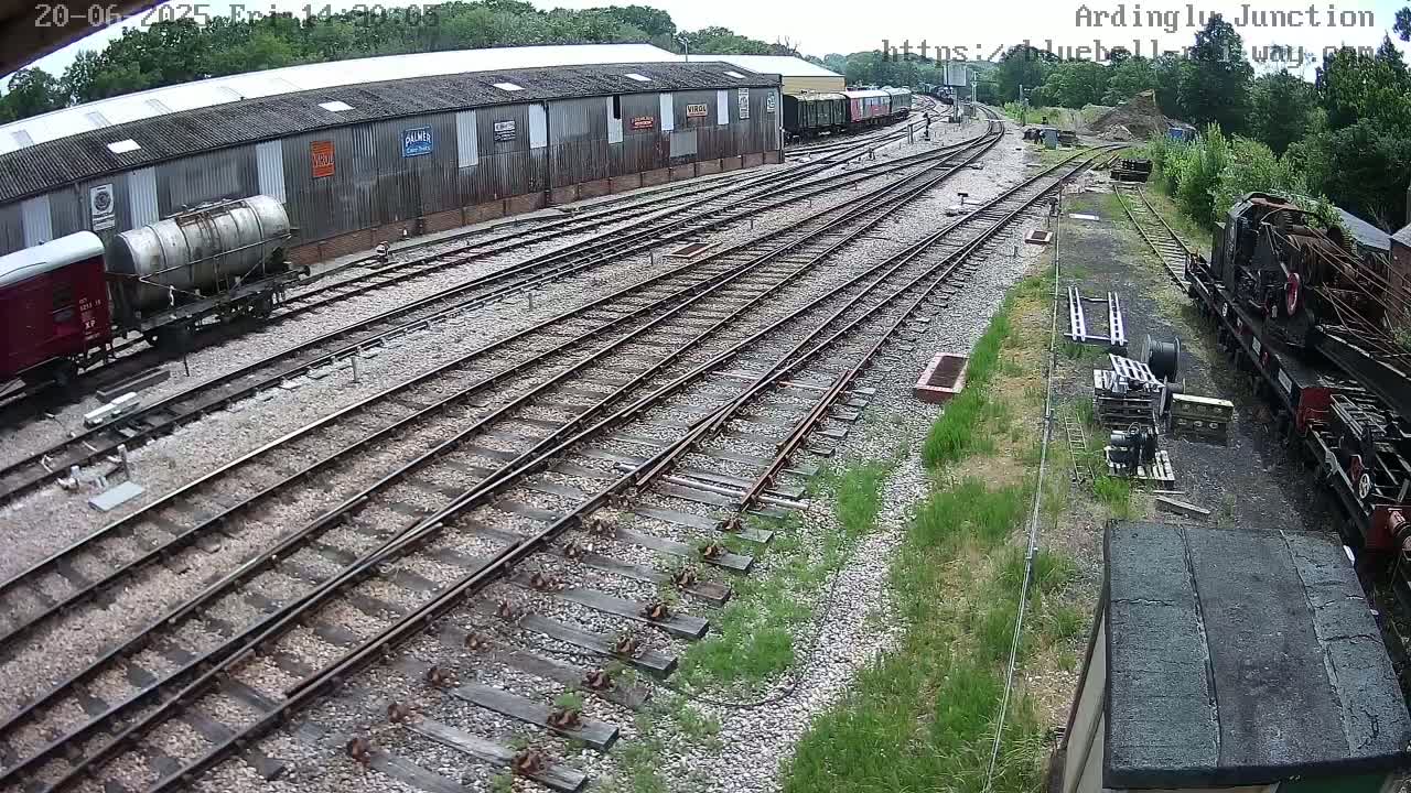 A high-angle view of a railway yard on a partly sunny day, showing multiple railway tracks branching out from a large building and several railway cars.