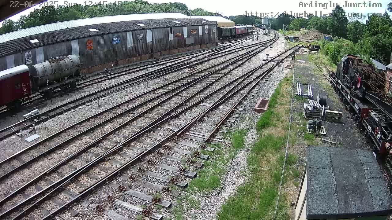 A railway yard with multiple train tracks, several rail cars, and a large industrial building is visible on a sunny day.