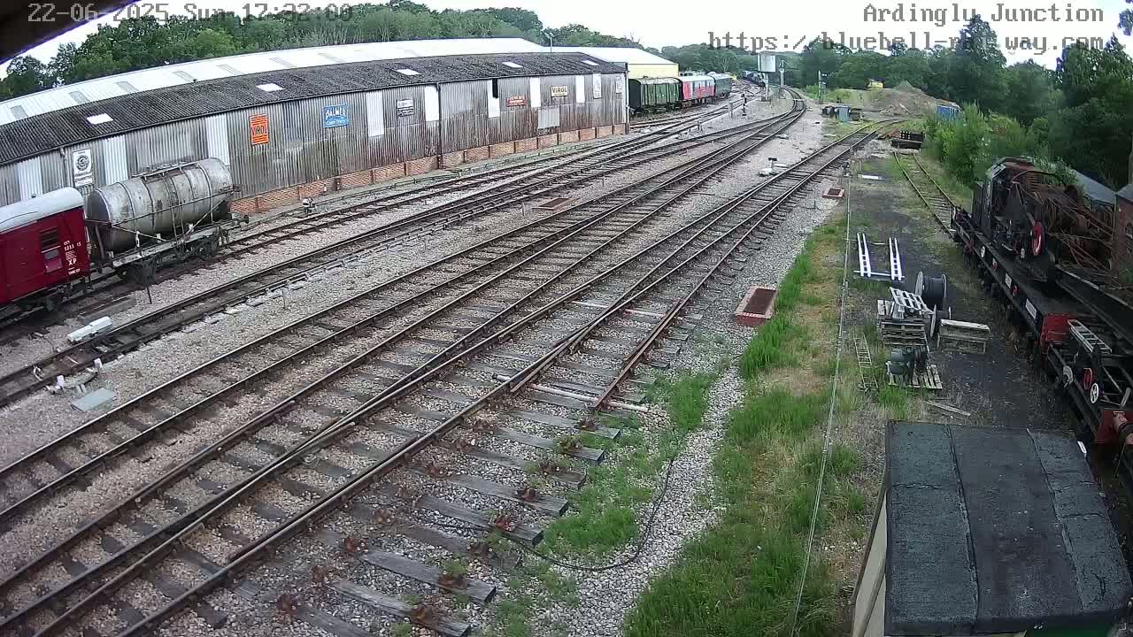 A railway yard with multiple train tracks, railway cars, and a large warehouse is visible under an overcast sky.