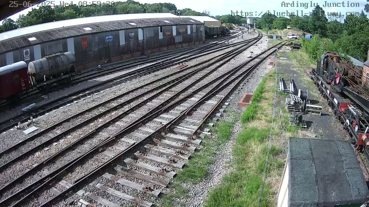 A high-angle view of a railway yard on a sunny day, showing multiple train tracks, railway cars, and a long building.