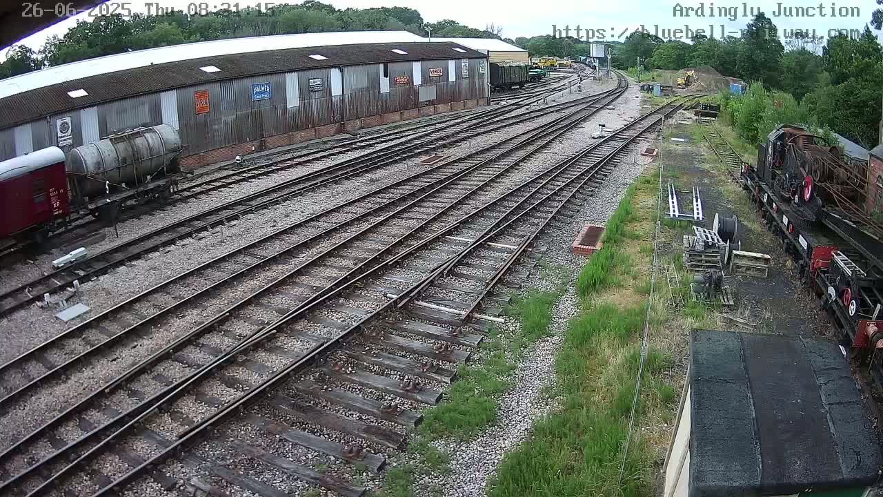 A high-angle view shows a railway yard with multiple tracks, railway cars, and a long building under a partly cloudy sky.