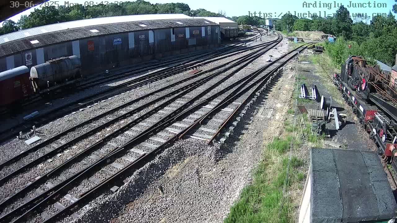 A sunny day at a railway yard shows numerous train tracks branching out from a long building with rail cars and a locomotive parked nearby.