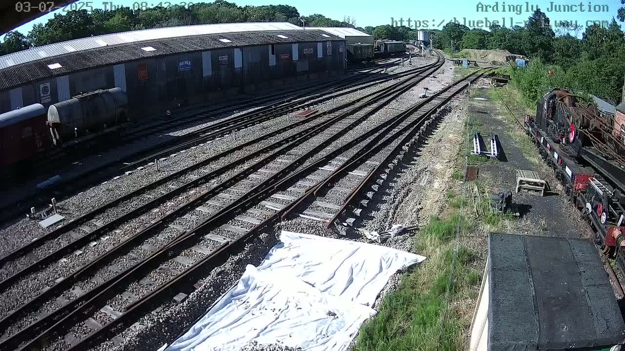 A sunny day at a railway yard shows multiple train tracks, railway cars, and a long shed.