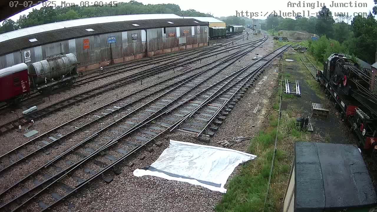 A railway yard with multiple train tracks, rail cars, and a large building, under an overcast sky.
