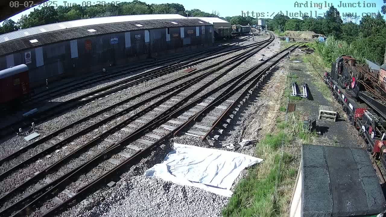 A sunny day at a railway yard shows multiple train tracks branching out from a long shed, with several train cars and a crane visible.