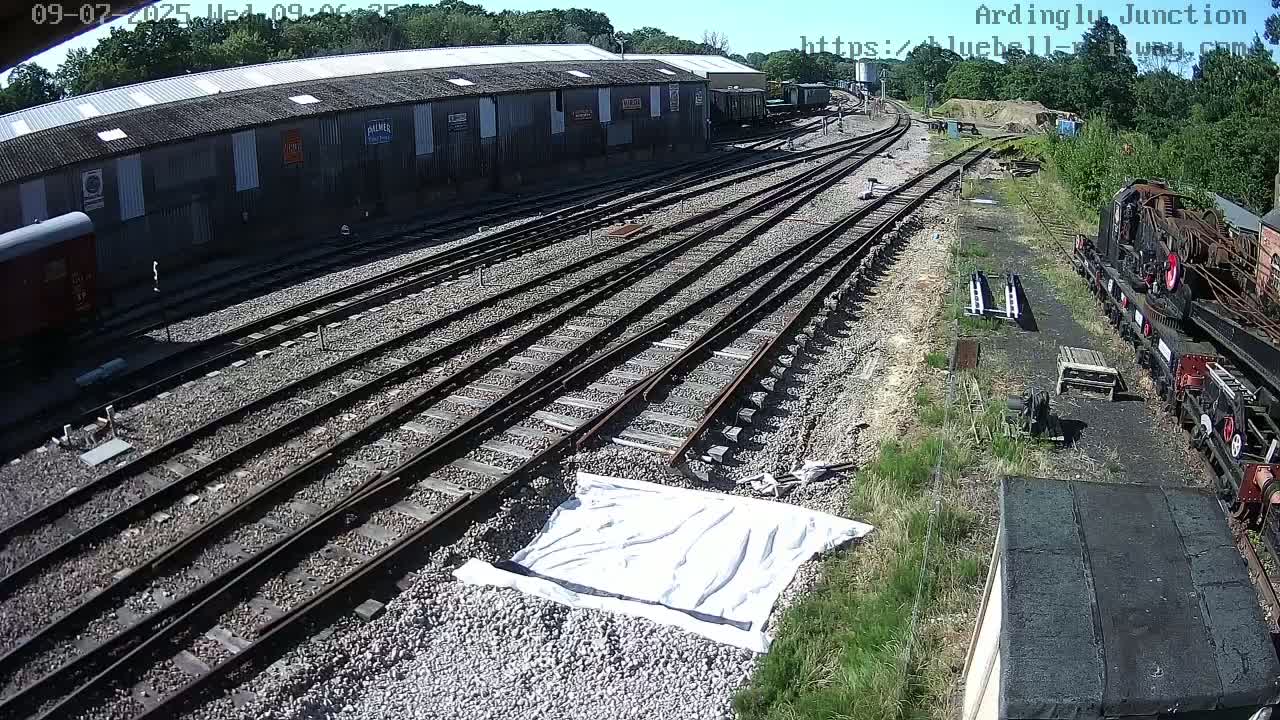 A sunny day at a railway yard shows multiple train tracks leading to a large building and a section of railway equipment.
