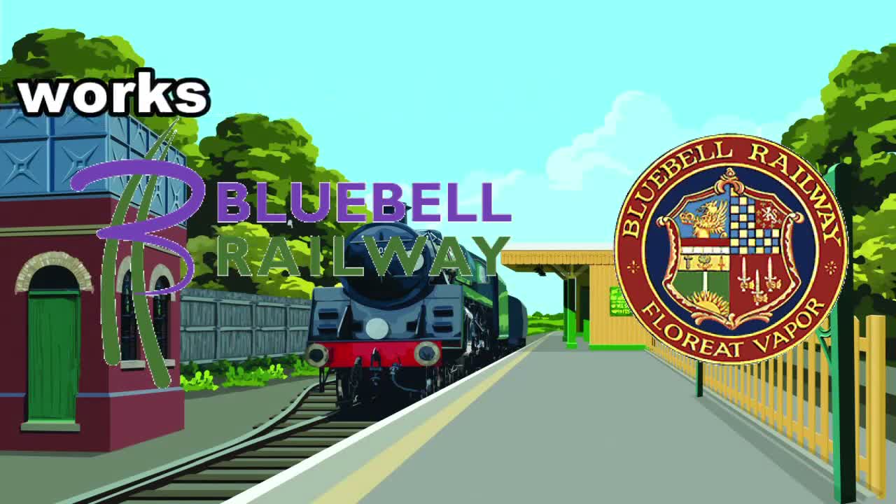 A steam locomotive is stopped at a sunny railway station platform with a red building to the left, green trees in the background, and a clear blue sky overhead.