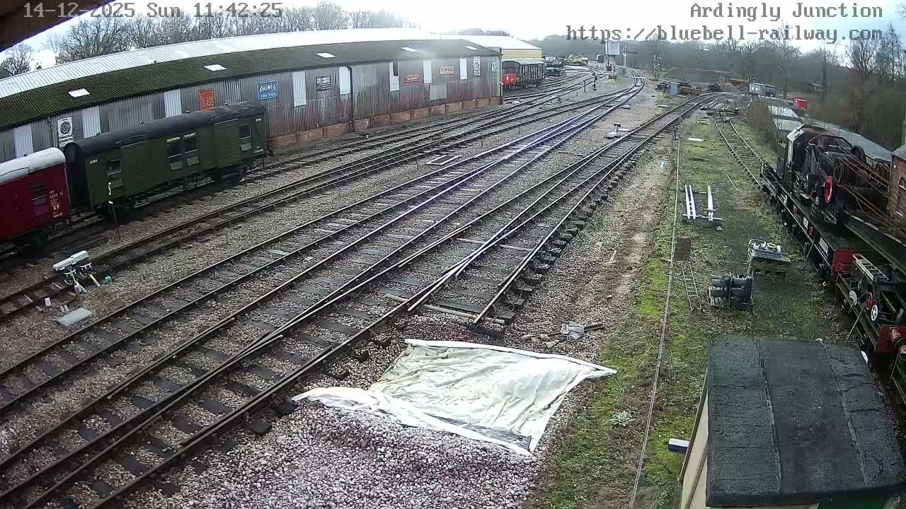 A steam locomotive is stopped at a sunny railway station platform with a red building to the left, green trees in the background, and a clear blue sky overhead.