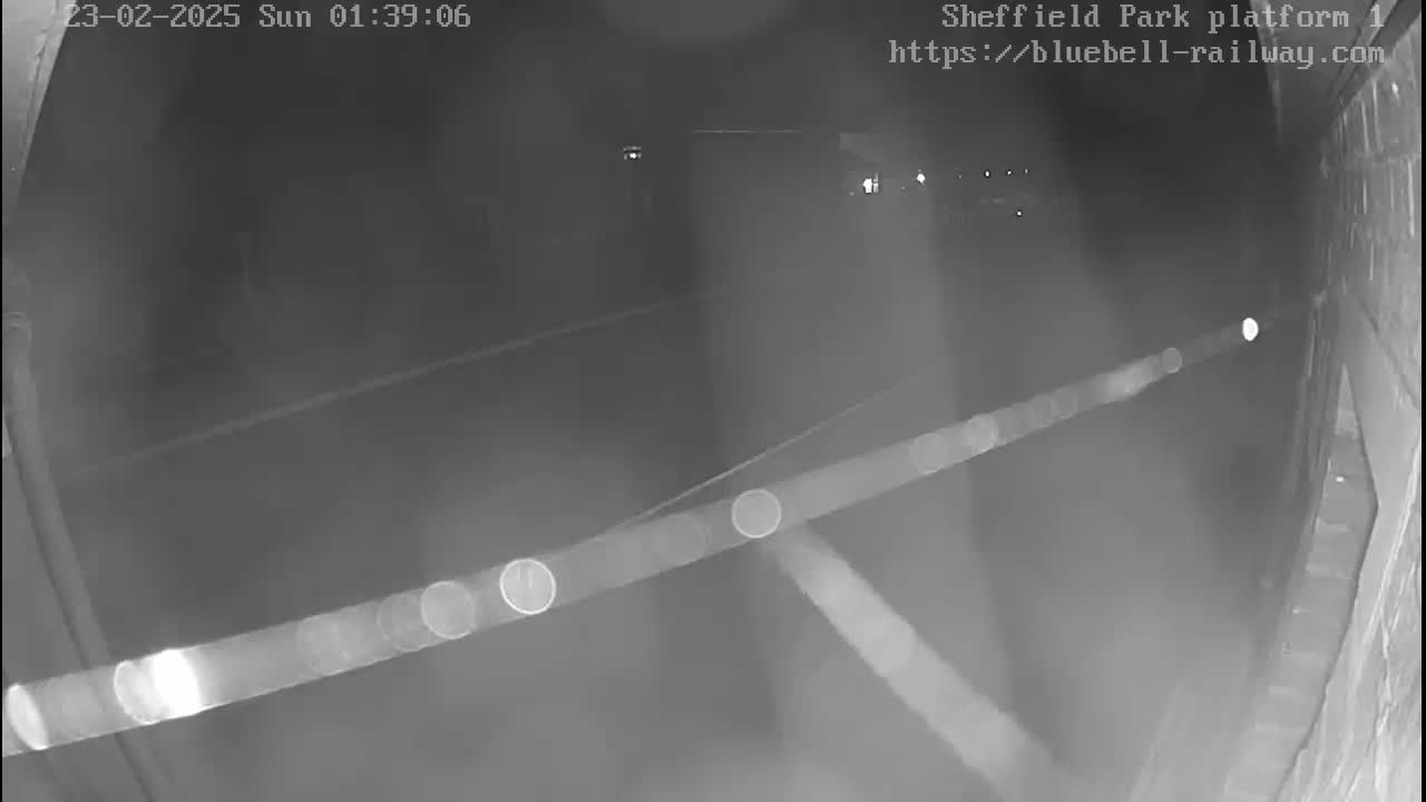 A nighttime, low-light security camera view shows a dark platform or roadway, possibly at a train station, with faint light sources visible in the distance.