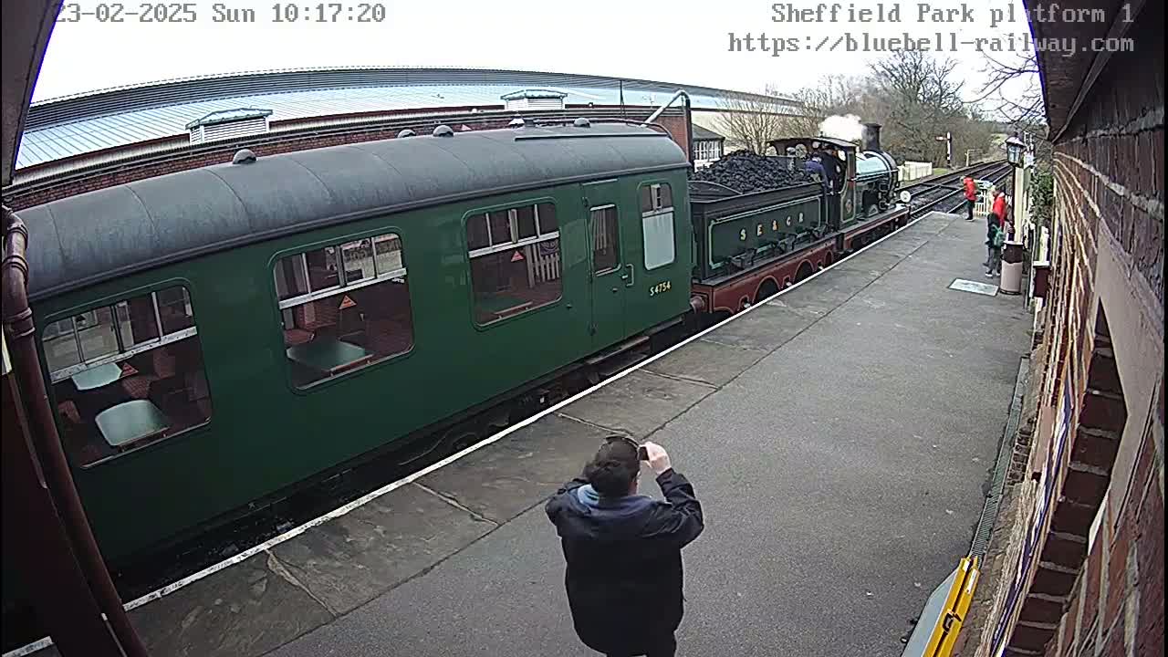 A green passenger train car is partially visible next to a steam locomotive at a train station on an overcast day.